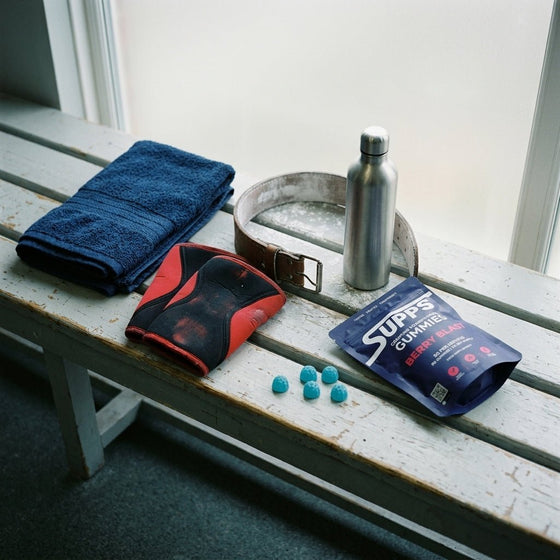 Supps Berry Blast creatine gummies pouch and loose gummies laid out on a changing-room bench with a towel, knee sleeves, weightlifting belt and water bottle